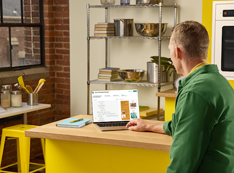Person in green shirt working at wooden desk with laptop in modern kitchen with yellow chairs and metal shelving