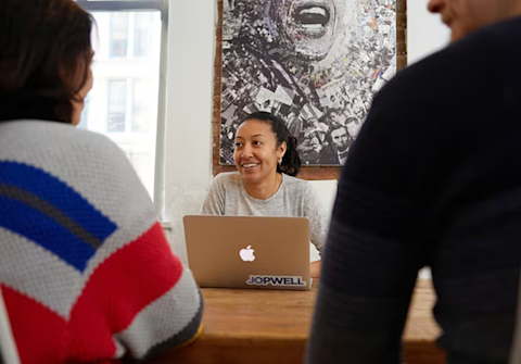 Professional woman smiling during a meeting at a wooden table, with a laptop and aerial map displayed on the wall behind her